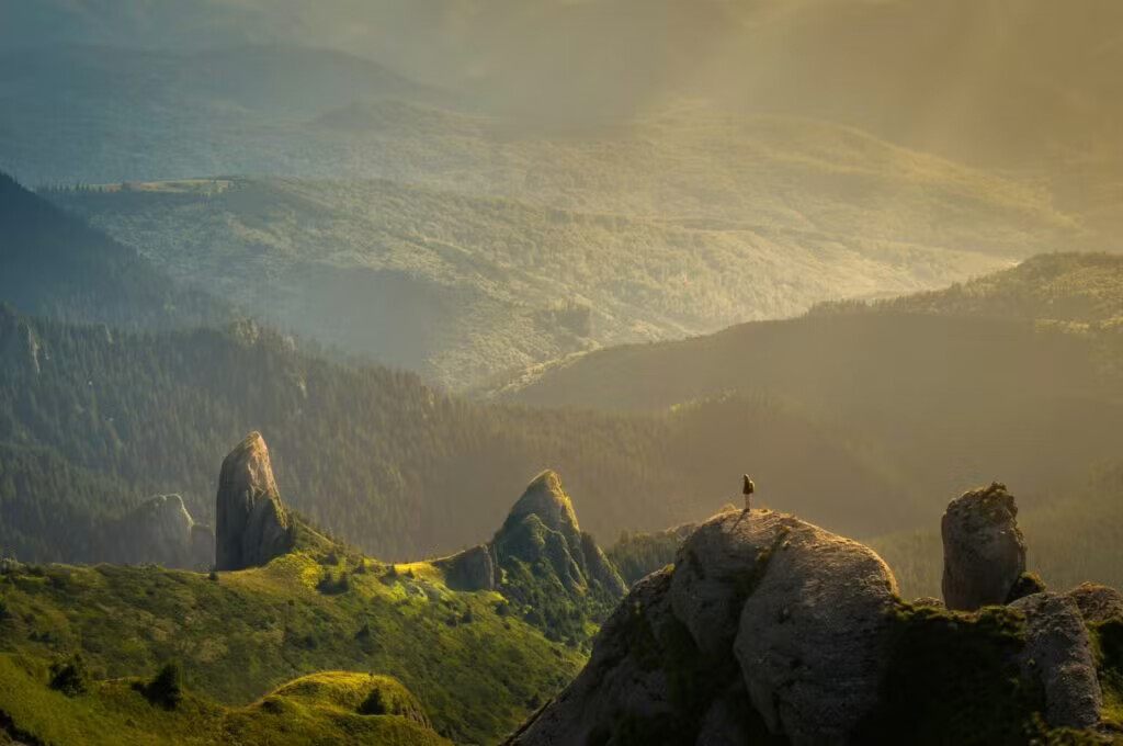 A person sitting on a rocky outcrop overlooking a mountainous landscape with a valley below, shrouded in mist.